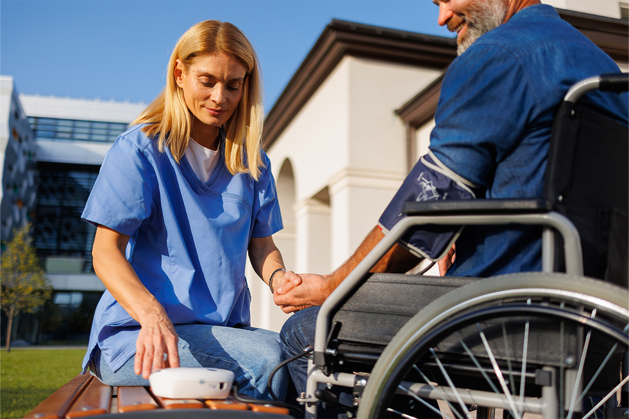 Female nurse in blue scrubs checking the blood pressure of a senior man in a wheelchair outdoors.