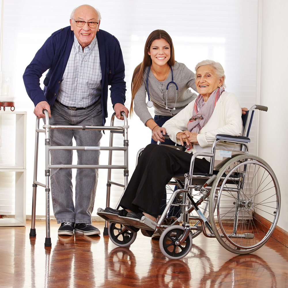 A smiling female caregiver stands between an elderly man with a walker and a woman in a wheelchair.